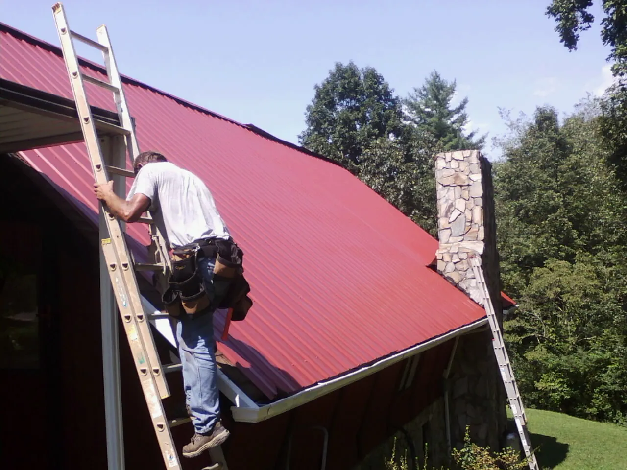 Roofer on a ladder beside a red metal roof and stone chimney — Soffit & Fascia Repair by Roofing Brothers in Weyers Cave